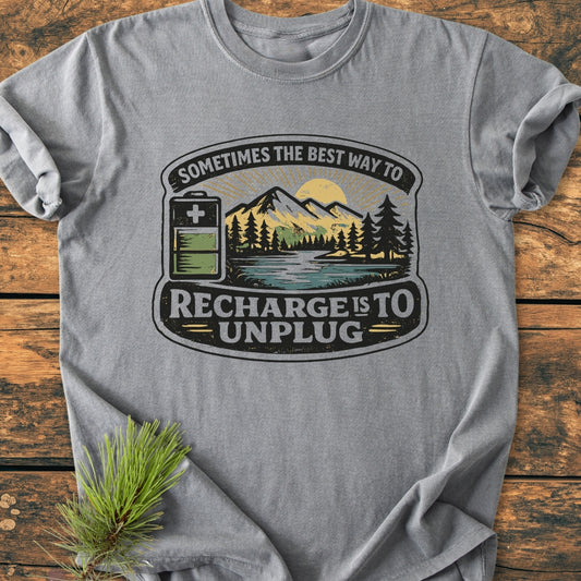 Unplug To Recharge - Vintage Wash Tee, featuring a heather-gray color with a mountain-and-lake graphic and slogan. Displayed flat with a pine sprig on a wood surface, highlighting its relaxed fit and durability.