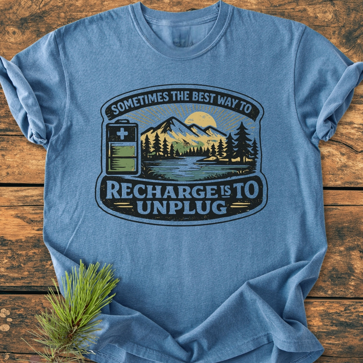 Unplug To Recharge - Vintage Wash Tee featuring a nature scene with mountains, sun, forest, lake, and battery icon, laid on wood. Enhanced by a pine bough, highlighting its rustic, cozy appeal.