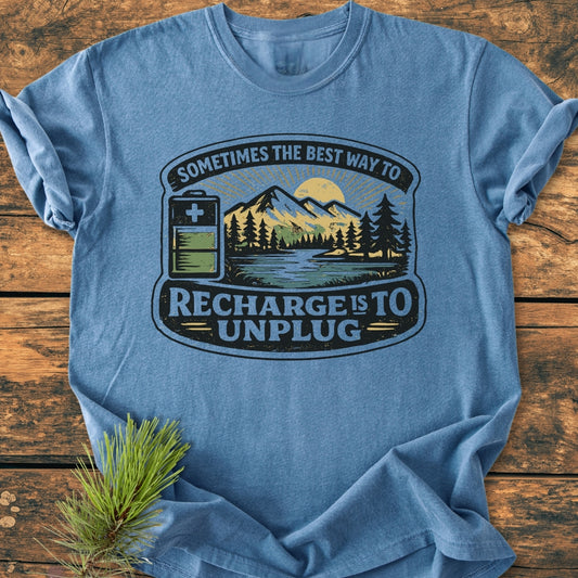 Unplug To Recharge - Vintage Wash Tee featuring a nature scene with mountains, sun, forest, lake, and battery icon, laid on wood. Enhanced by a pine bough, highlighting its rustic, cozy appeal.