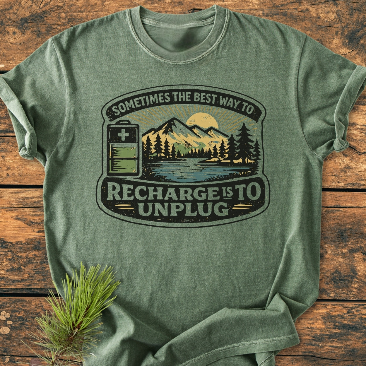 Unplug To Recharge - Vintage Wash Tee features a scenic print with pine trees, mountains, and a battery icon, emphasizing relaxation. Laid on a rustic surface, highlighting its relaxed fit and soft texture.