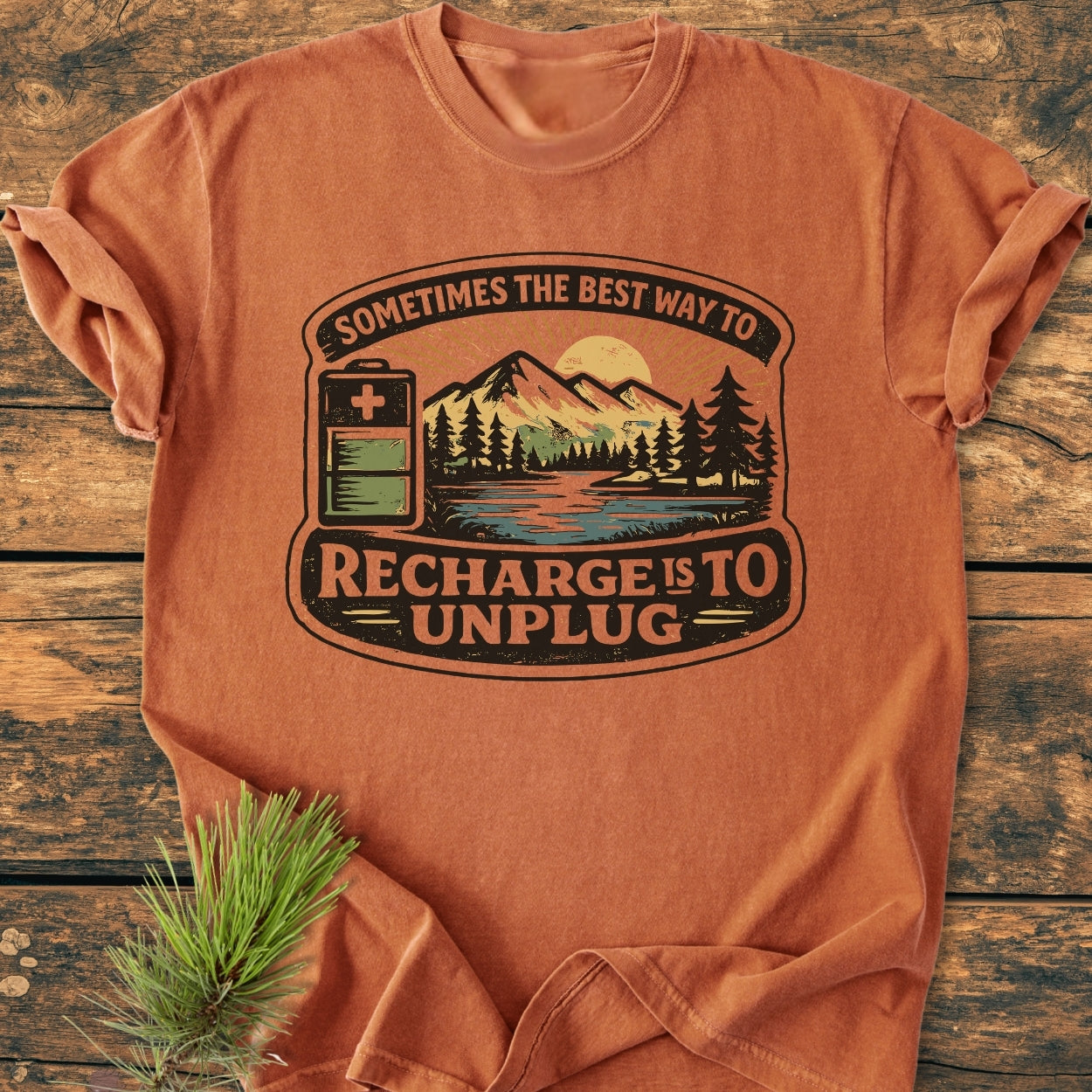 Unplug To Recharge - Vintage Wash Tee: Burnt-orange short-sleeve with forest-and-mountain graphic, 'Recharge is to unplug' text. Displayed on wood, featuring soft-washed, garment-dyed cotton for a relaxed fit.