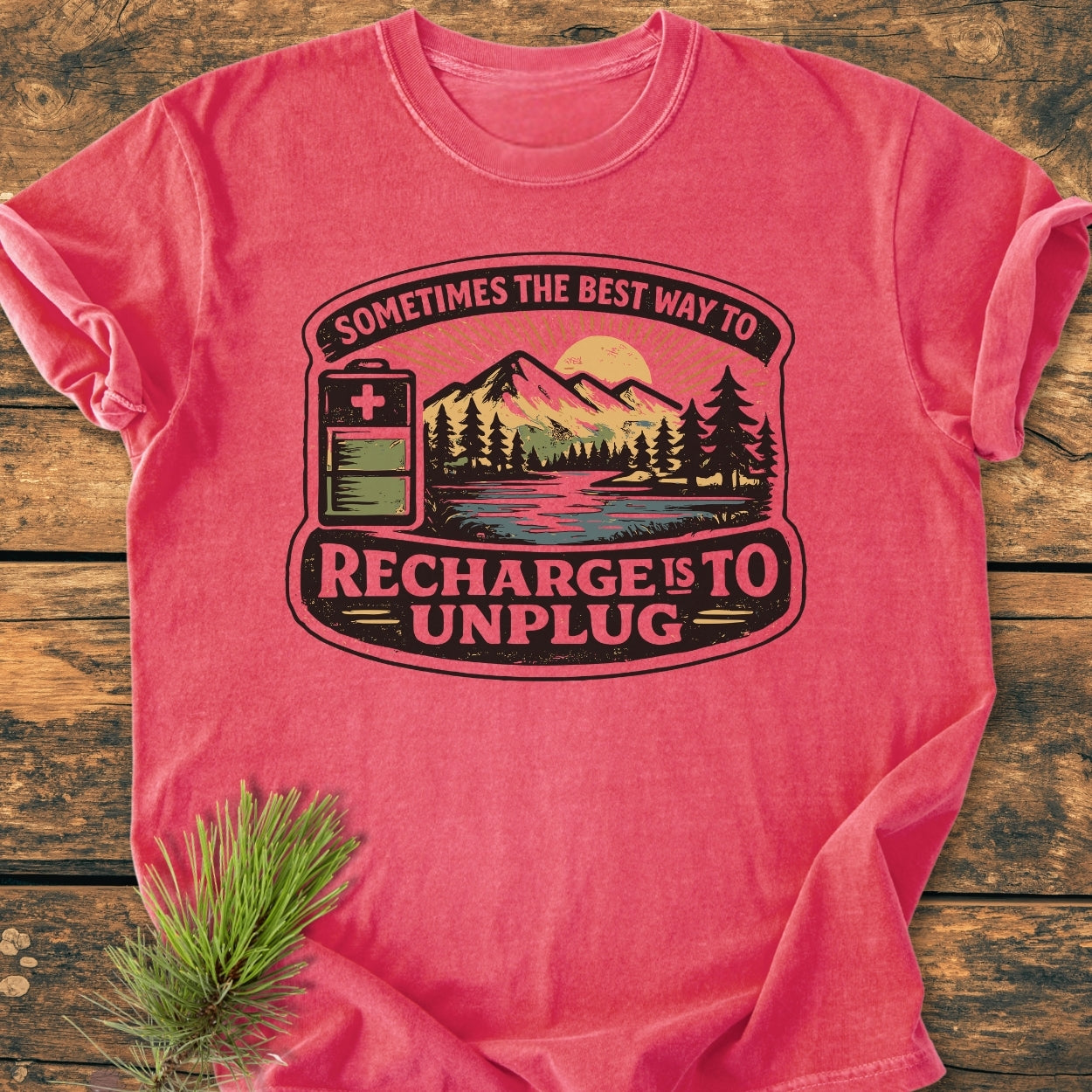 Unplug To Recharge - Vintage Wash Tee displays a mountain lake scene with a battery icon and slogan. Laid on wood, it showcases the Comfort Colors' garment-dyed, relaxed fit, 100% cotton fabric.