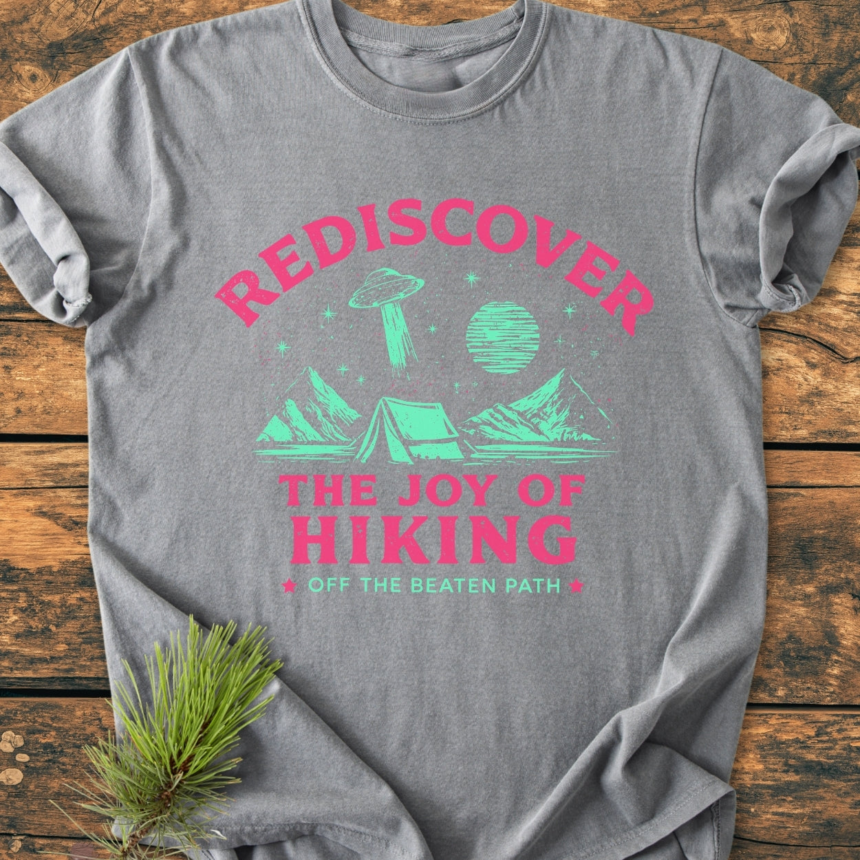 Rediscover The Joy Of Hiking - Vintage Wash Tee features a whimsical outdoor scene with mountains, a tent, stars, and a flying saucer, printed in bright colors on a heather-gray background.