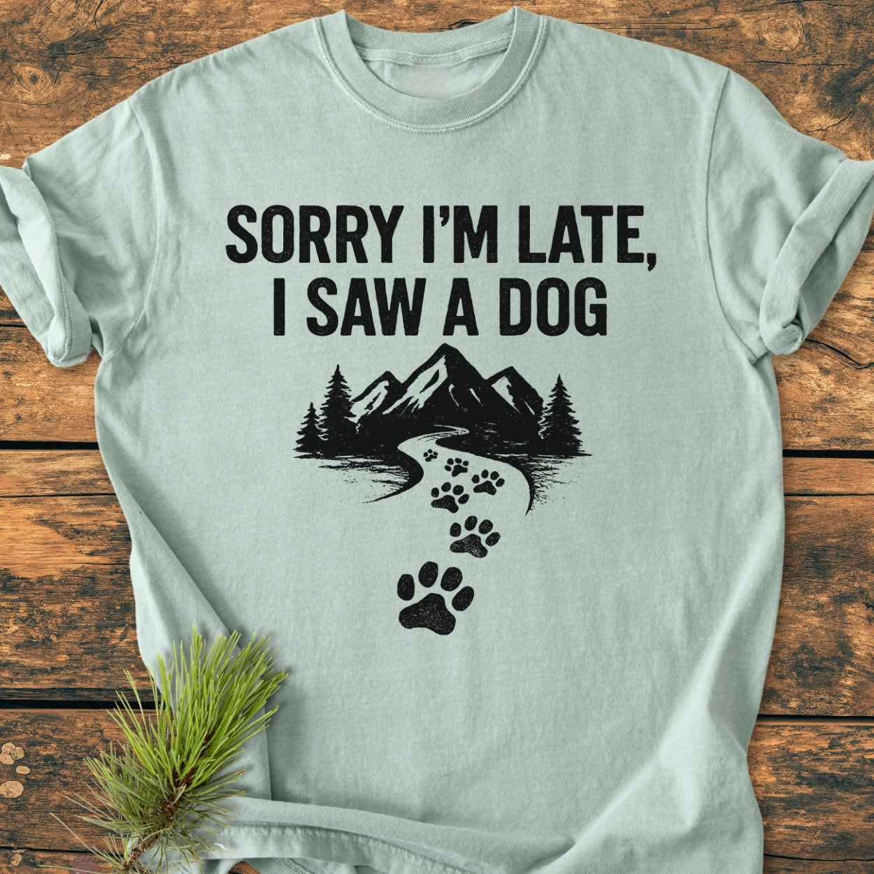Sorry I'm Late - Vintage Wash Tee with bold lettering and mountain scene, featuring durable, soft-washed fabric, laid on wooden surface with pine sprig accent.