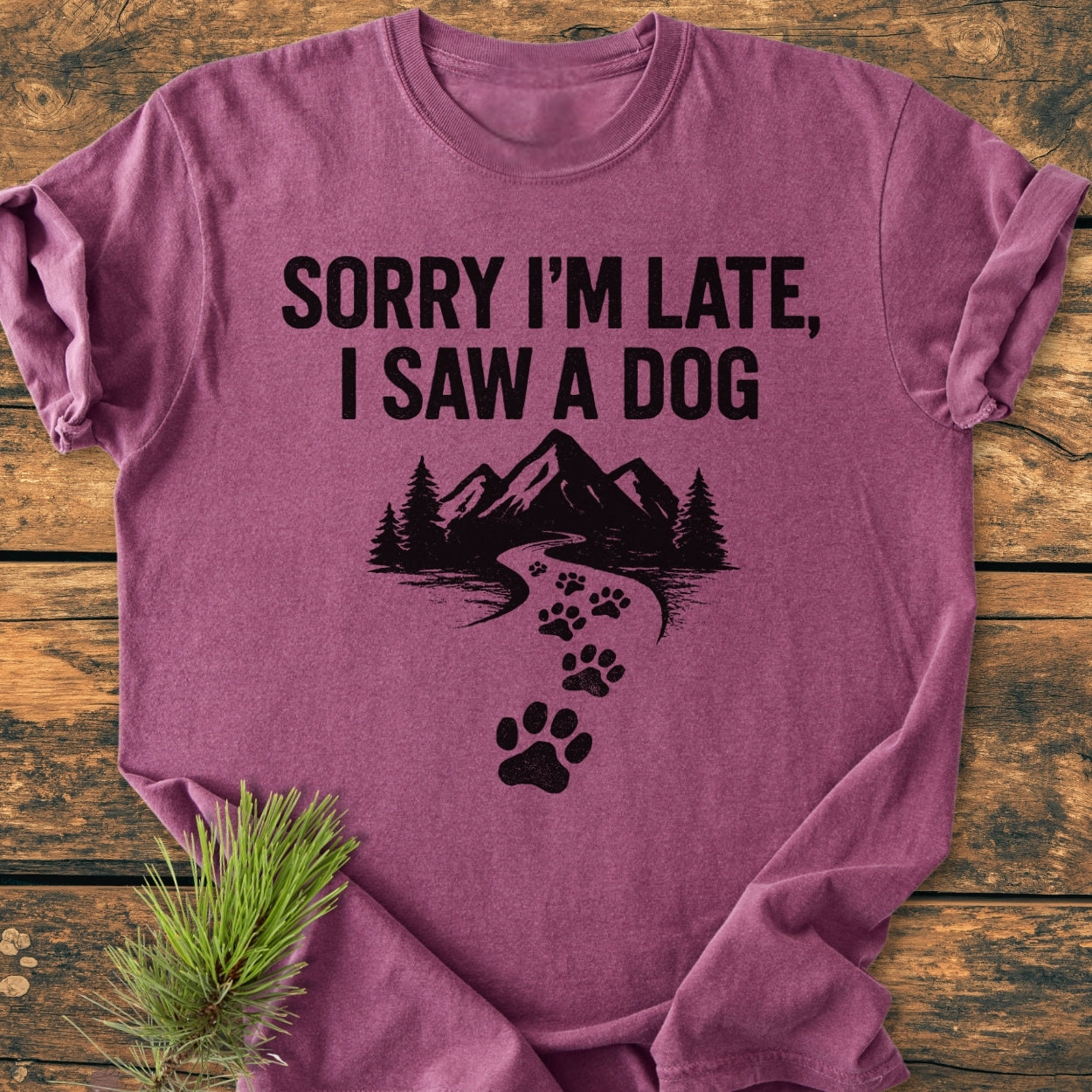 Sorry I'm Late - Vintage Wash Tee, featuring bold lettering and a mountain scene with paw prints on a heathered purple T-shirt, laid on wood with a pine sprig for a rustic touch.