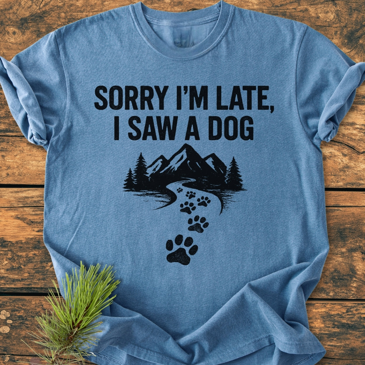 Sorry I'm Late - Vintage Wash Tee featuring a mountain-and-forest design and dog paw trail, laid flat on wood with pine sprig, showcasing its soft, relaxed fit and durable stitching.