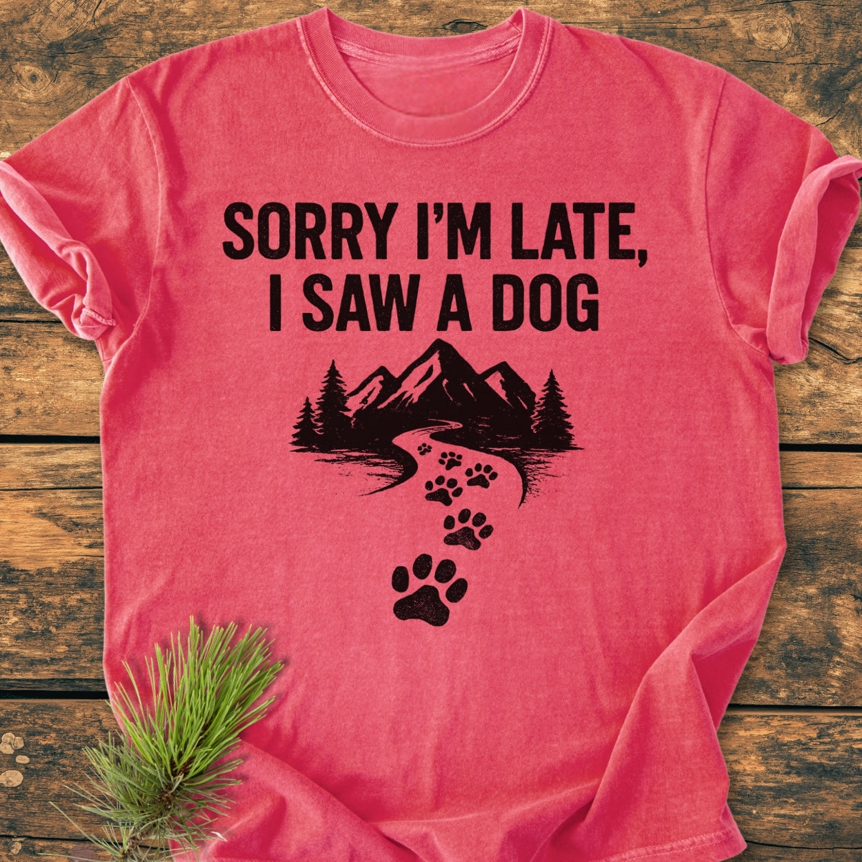 Sorry I'm Late - Vintage Wash Tee features a black graphic with 'SORRY I’M LATE, I SAW A DOG' text, mountain scene, and paw-print path, laid on wooden surface with pine sprig.