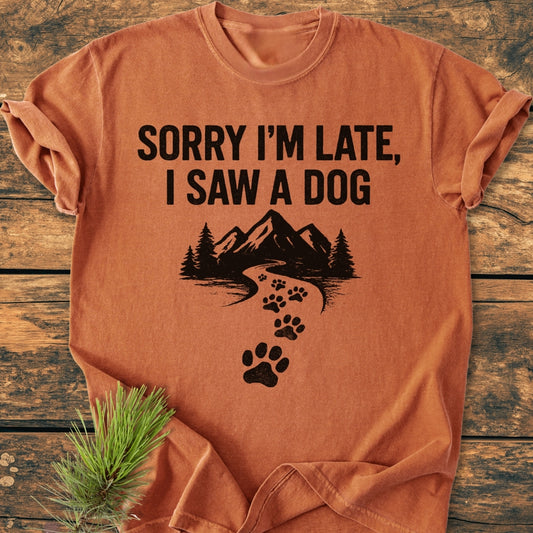 Sorry I'm Late - Vintage Wash Tee features bold text and a trail graphic with paw prints, laid on wood with an evergreen sprig, emphasizing its relaxed fit and cozy, garment-dyed fabric.