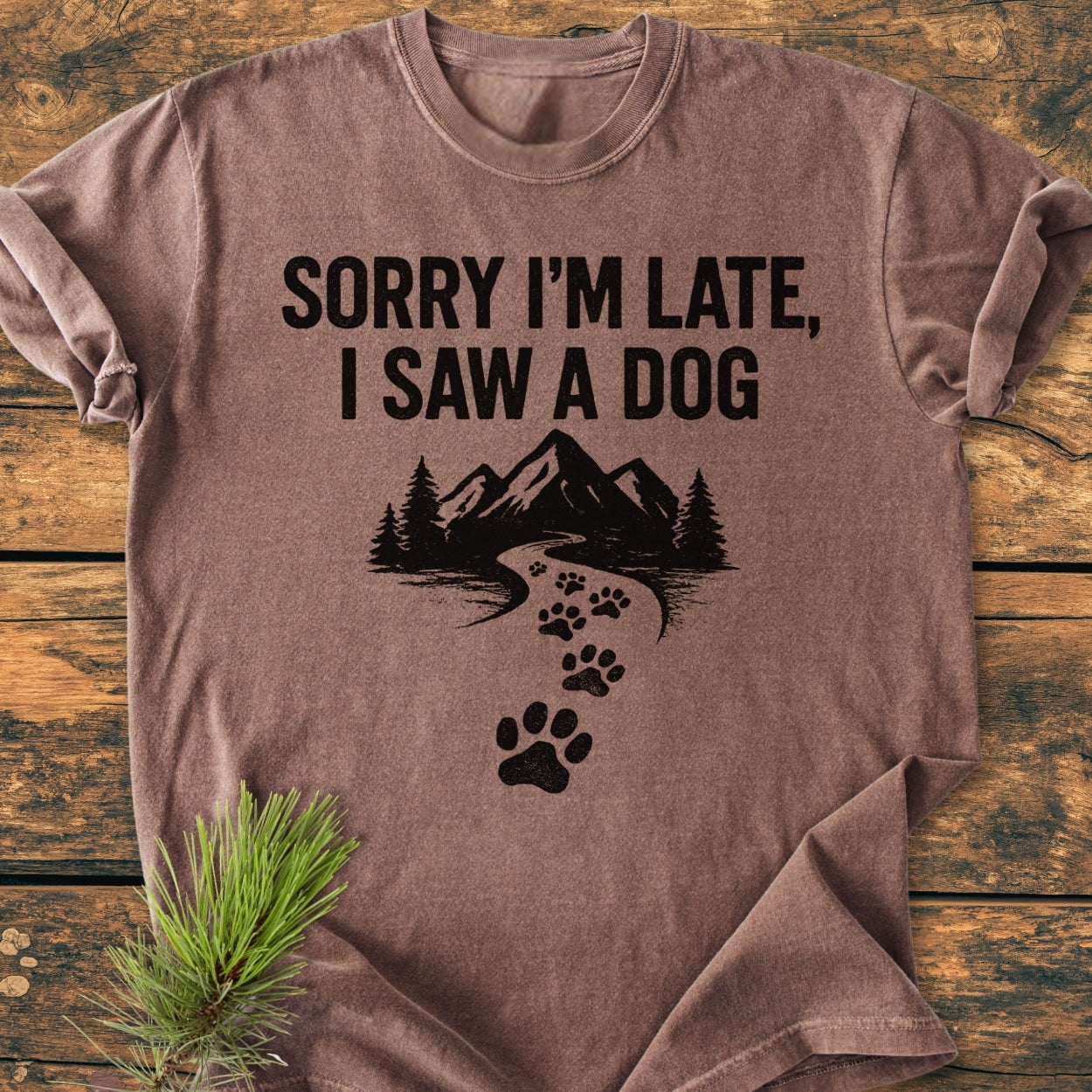 Sorry I'm Late - Vintage Wash Tee: A brown short-sleeve T-shirt with bold text and a mountain graphic. Laid flat on wood, featuring soft-washed, 100% ring-spun cotton for a cozy, relaxed fit.