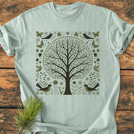 Nature Love - Vintage Wash Tee features a sea-foam green shade with a large, stylized “tree of life” design, adorned with birds and flowers, laid flat on a wooden surface.
