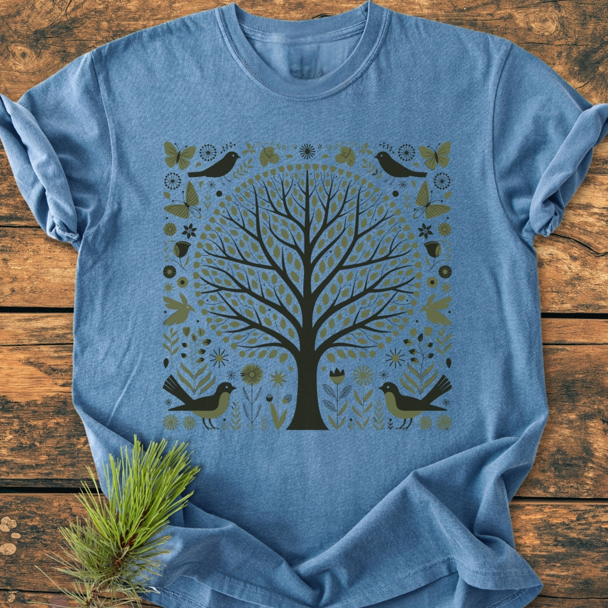 Nature Love - Vintage Wash Tee: A blue, short-sleeved tee featuring a stylized tree graphic with birds and butterflies, laid on a wooden surface with a pine sprig beside it.
