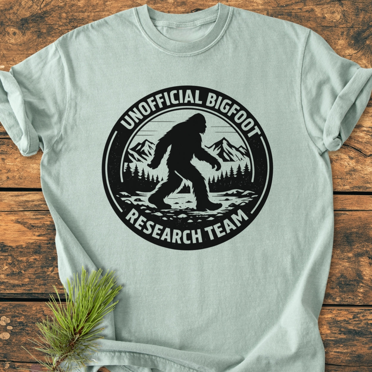 Unofficial Bigfoot Research Team - Vintage Wash Tee featuring Bigfoot silhouette against mountains, laid on wooden surface with pine branch, highlighting garment-dyed cotton's softness and durable, relaxed fit.