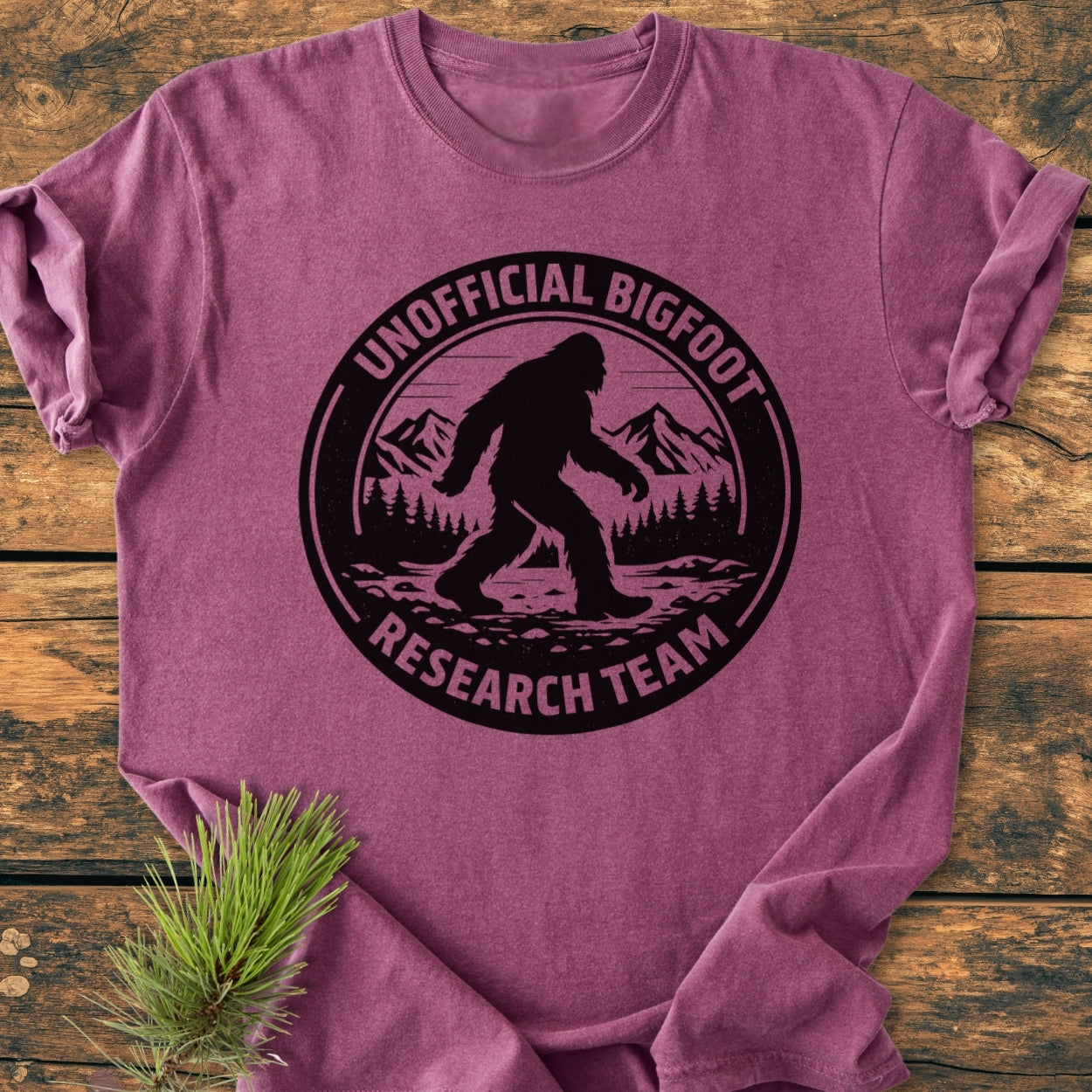 Unofficial Bigfoot Research Team - Vintage Wash Tee featuring a black Bigfoot silhouette graphic against pines and mountains on a mauve tee, styled with an evergreen branch on a wooden surface.