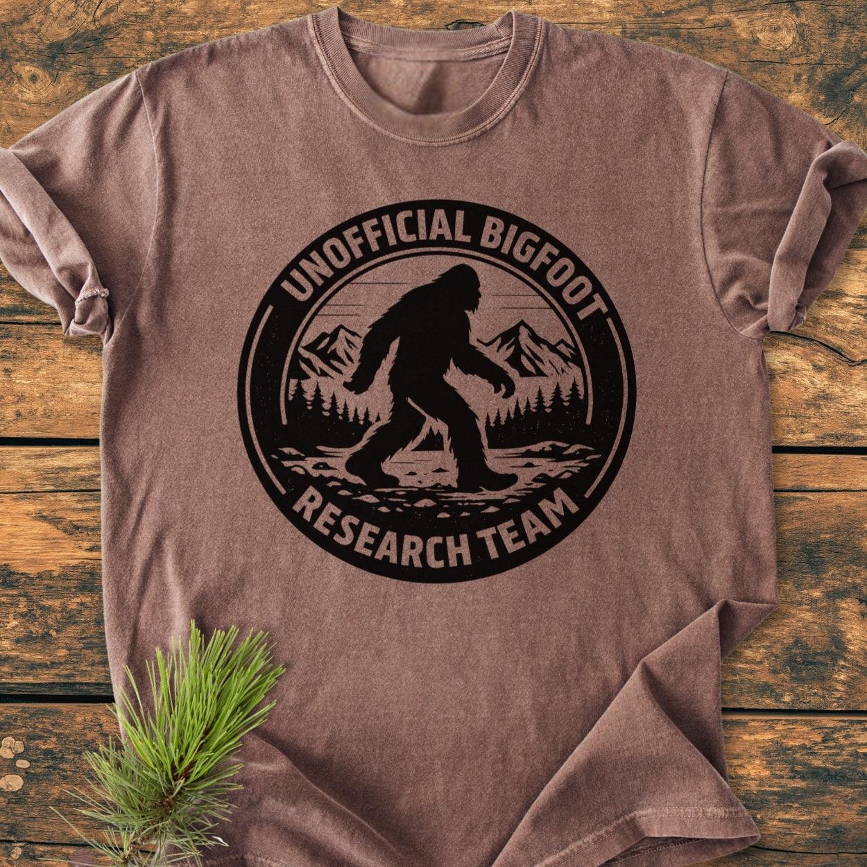 Unofficial Bigfoot Research Team - Vintage Wash Tee features a Bigfoot graphic with pine trees and mountains, laid flat on wood. Made of soft, durable, 100% ring-spun cotton for everyday comfort.