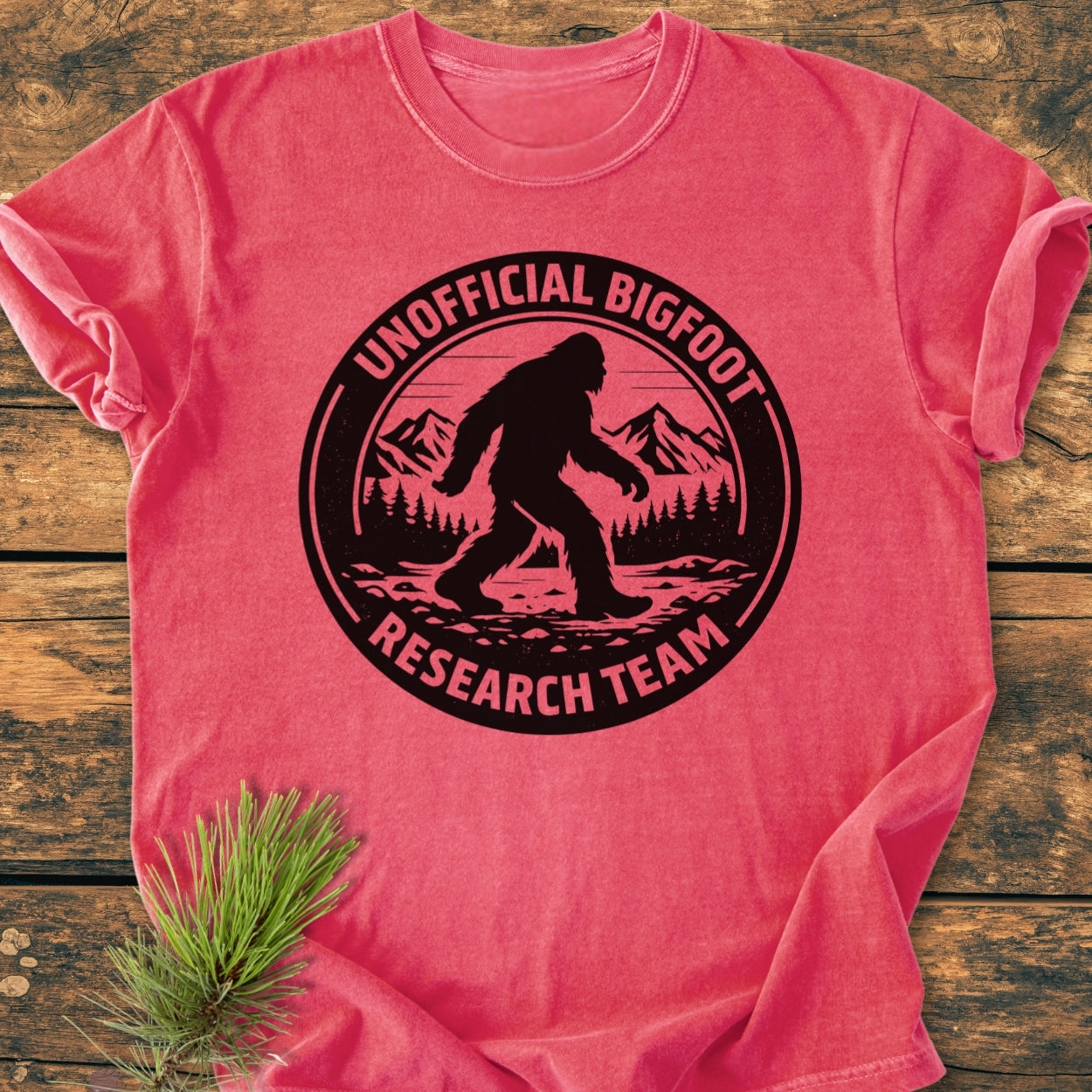 Unofficial Bigfoot Research Team - Vintage Wash Tee features a black Bigfoot graphic on a faded red shirt, lying on wood. The tee combines comfort with a whimsical design, ideal for everyday wear.
