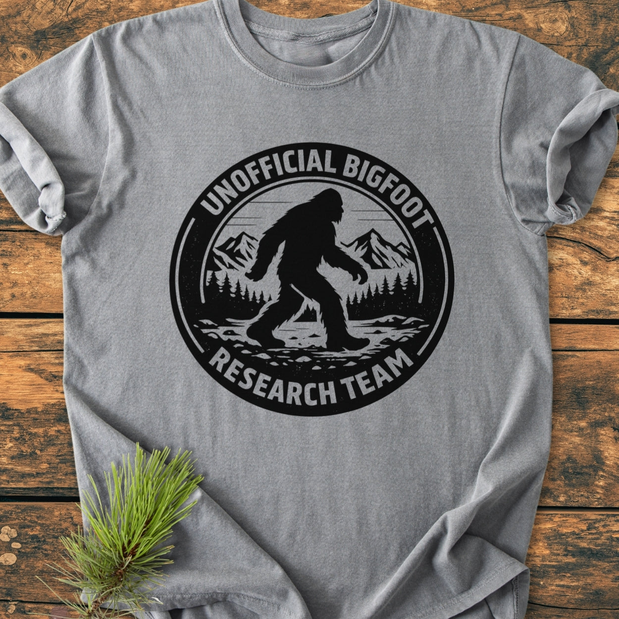 Unofficial Bigfoot Research Team - Vintage Wash Tee featuring a black Bigfoot graphic and text on a gray shirt, laid on wood, with a pine branch beside it.