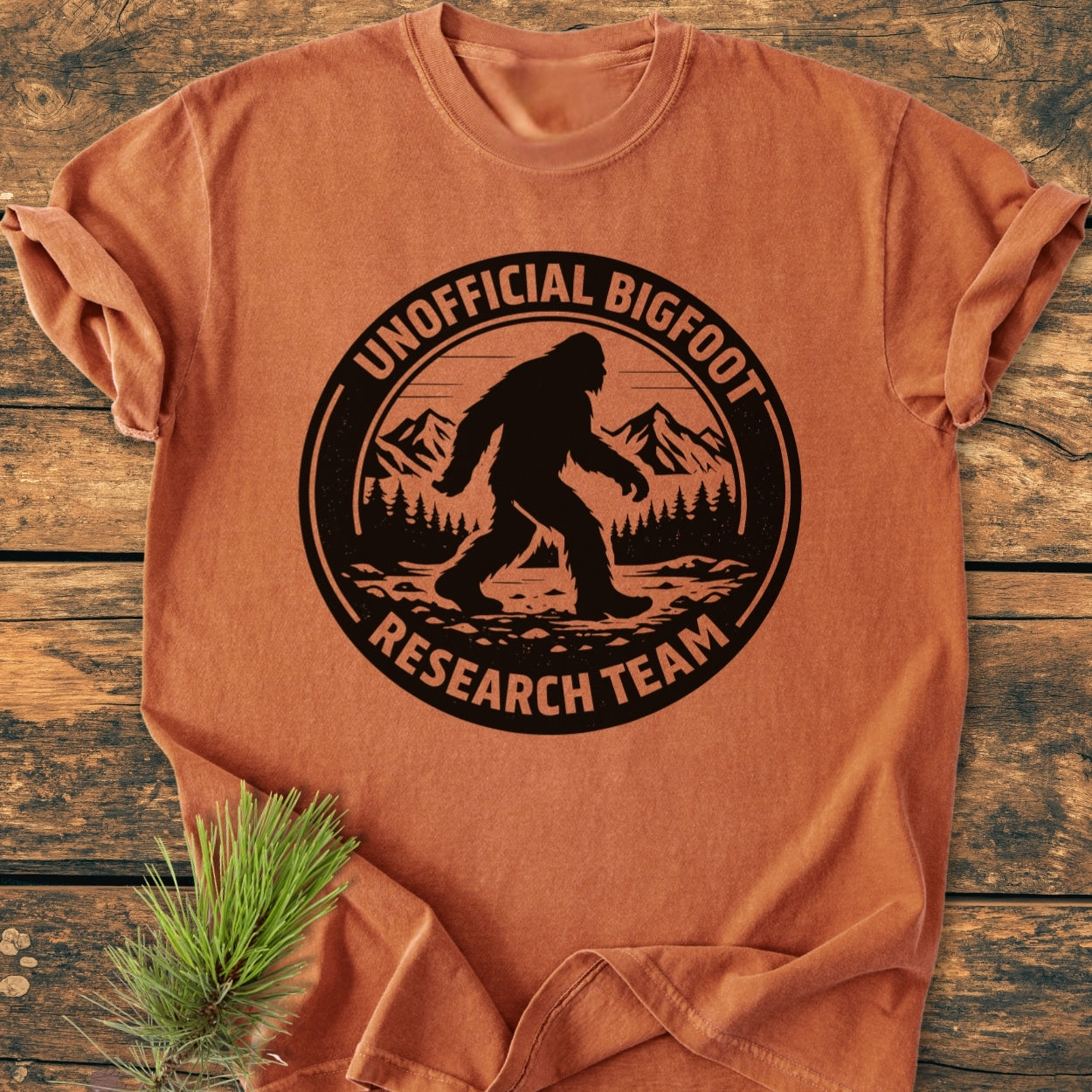 Unofficial Bigfoot Research Team - Vintage Wash Tee, folded, features a large black Bigfoot silhouette graphic with text, on a burnt-orange background, displayed on a wooden surface with a pine branch accent.