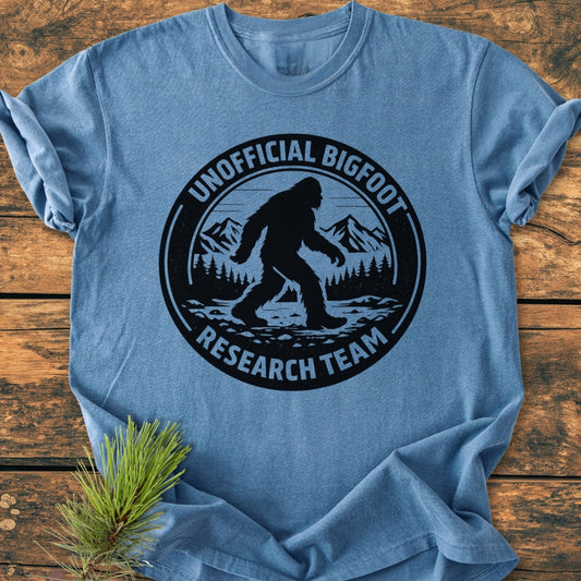 Unofficial Bigfoot Research Team - Vintage Wash Tee: Blue T-shirt with a black Bigfoot graphic and text on front, laid on wood surface with decorative pine sprig. 100% ring-spun cotton, relaxed fit.