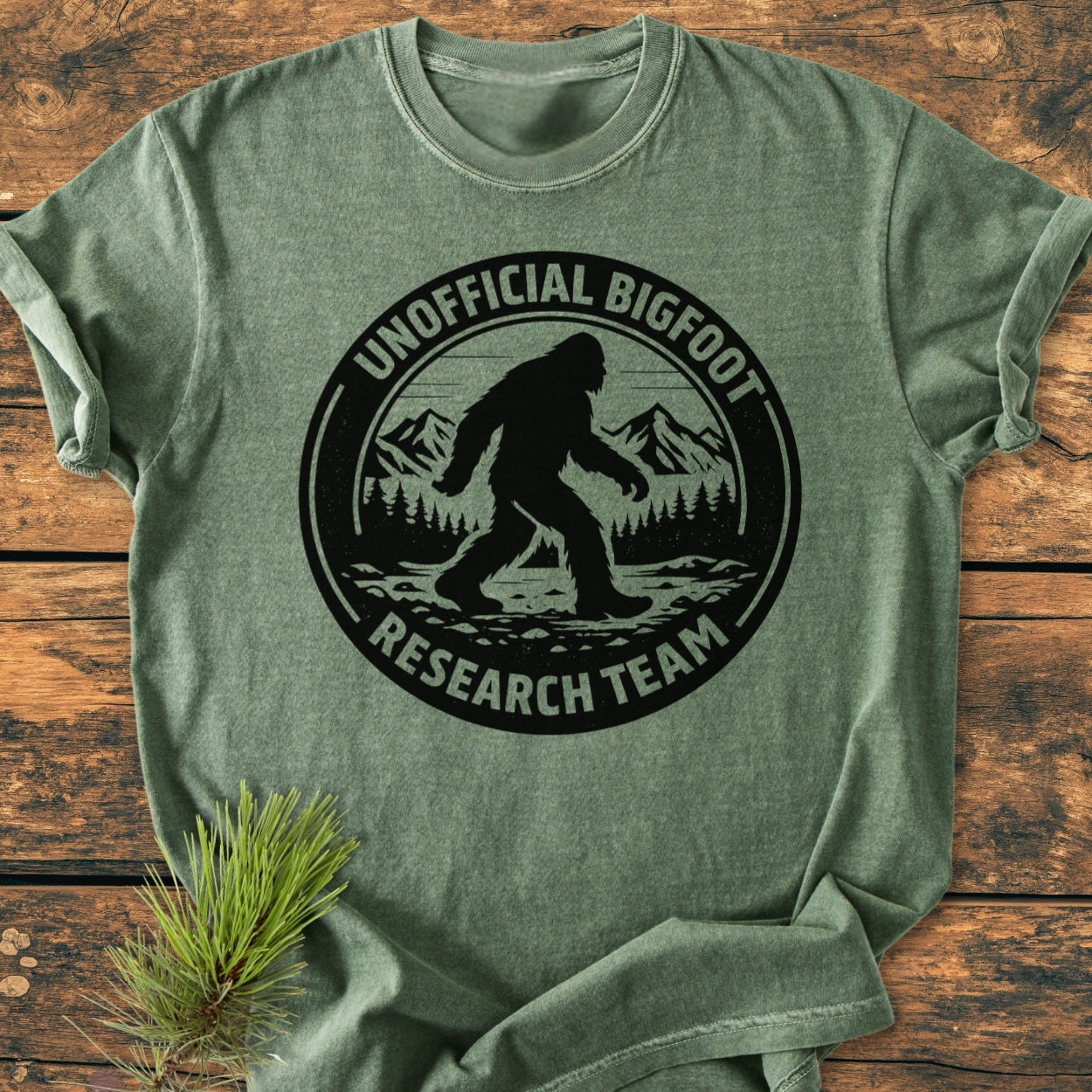 Unofficial Bigfoot Research Team - Vintage Wash Tee: A green T-shirt featuring a black emblem with Bigfoot's silhouette against a mountain and forest, laid on a wooden surface.