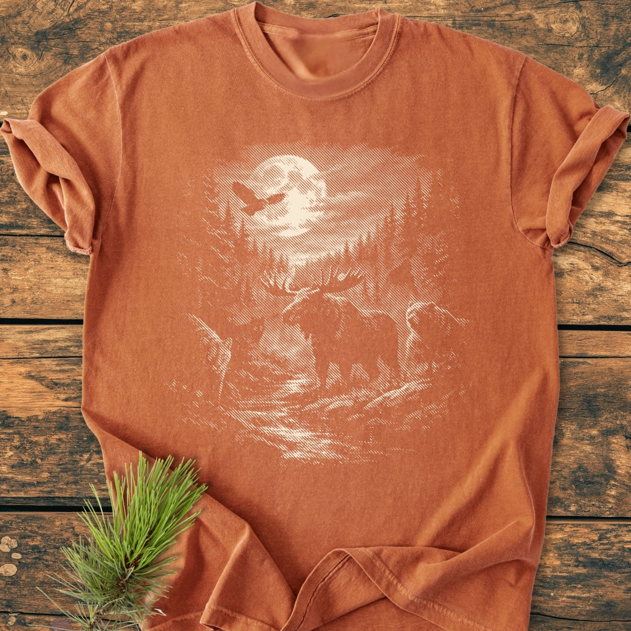 Moonlit Woodland - Vintage Wash Tee features a sketch-style forest scene with a moose and owl on a burnt-orange T-shirt, displayed flat on a rustic wooden surface with a pine sprig accent.