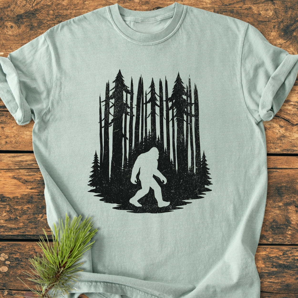 Nighttime Wanderer - Vintage Wash Tee features a stylized forest scene with a Sasquatch silhouette, screen-printed on a pale-green shirt. Laid on wood, accompanied by a decorative pine sprig.