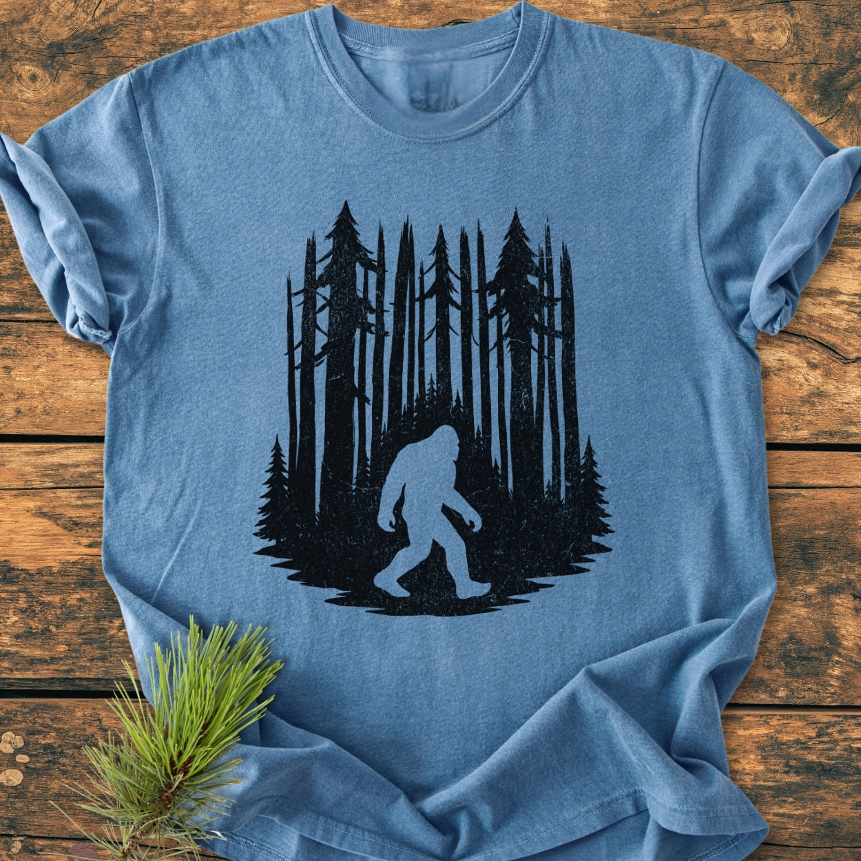 Nighttime Wanderer - Vintage Wash Tee features Bigfoot silhouette against pine trees, laid flat with a pine twig accent on a wooden background. Crafted from soft, durable 100% ring-spun cotton.