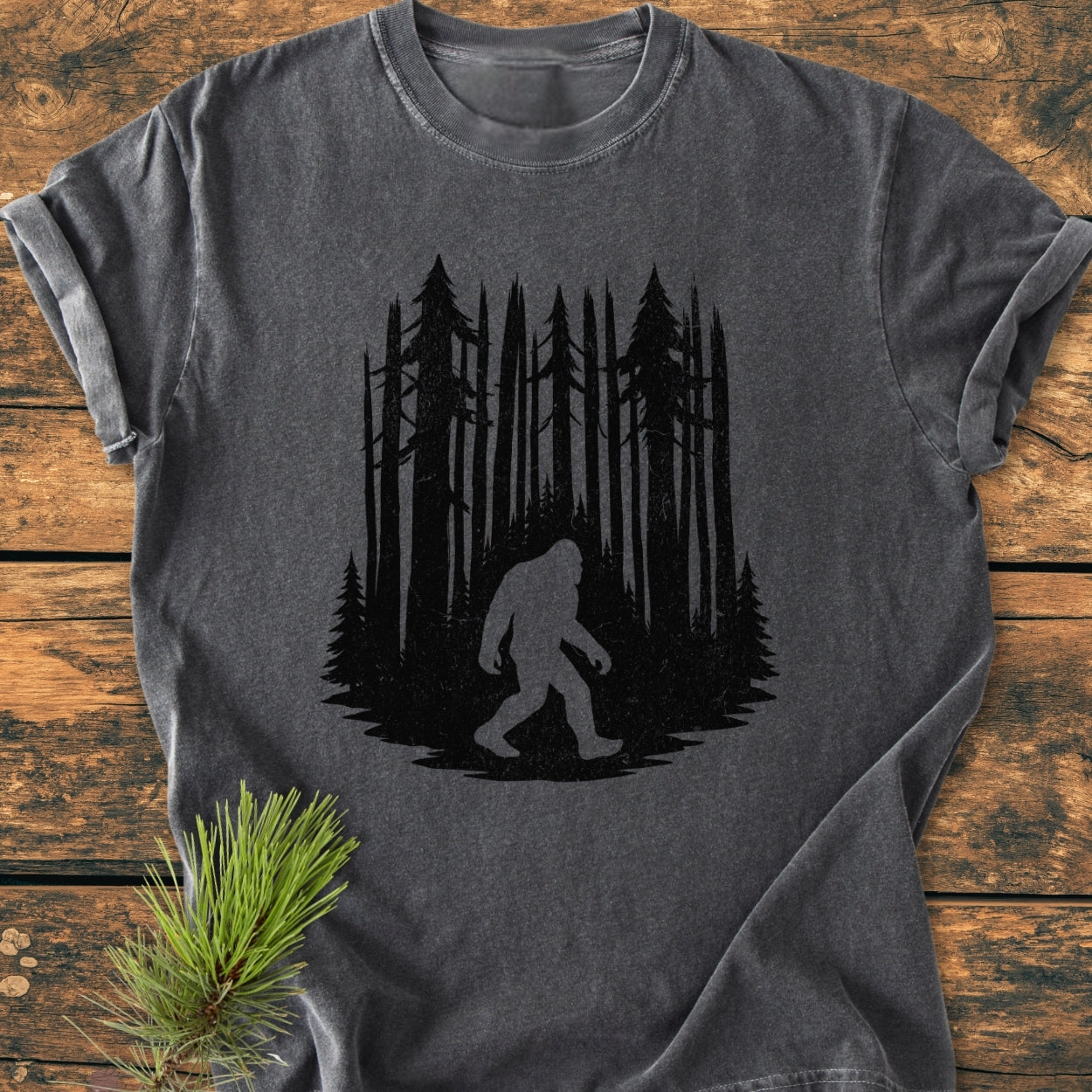 Nighttime Wanderer - Vintage Wash Tee: A charcoal-gray T-shirt featuring a black silhouette of a dense pine forest with a classic Sasquatch figure, laid on a wooden tabletop with a pine sprig beside it.