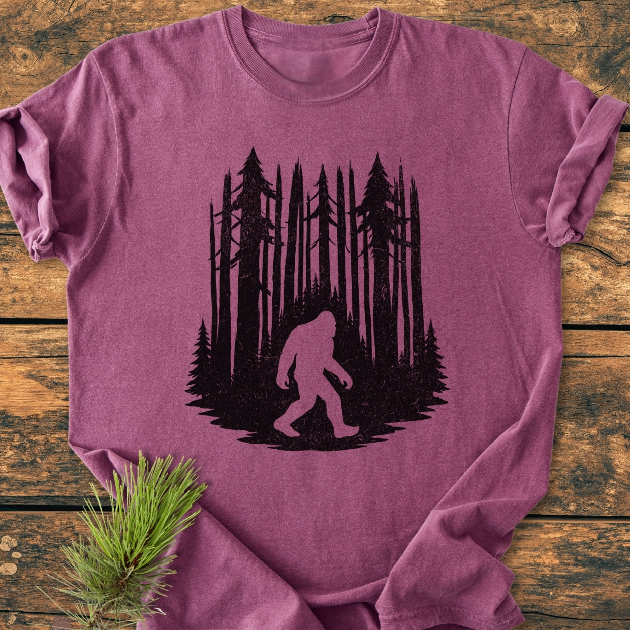 Nighttime Wanderer - Vintage Wash Tee features a black Bigfoot silhouette and pine trees printed on a heathered plum T-shirt, displayed flat with a pine branch. Ideal for everyday coziness.