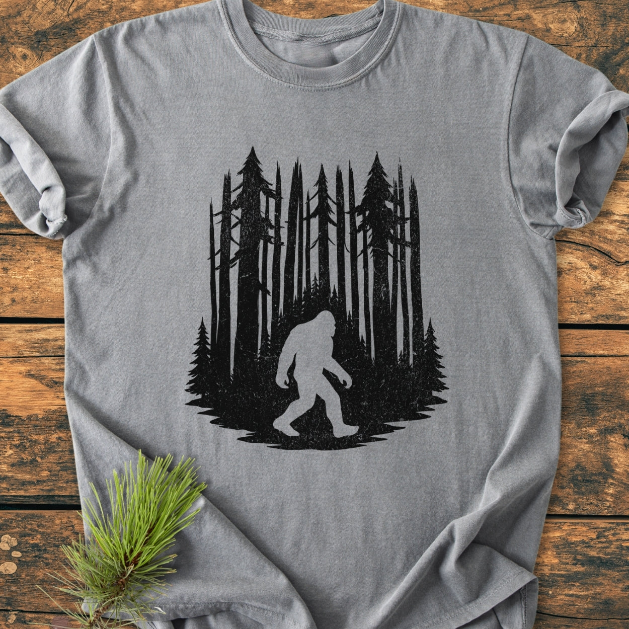 Nighttime Wanderer - Vintage Wash Tee features a Bigfoot silhouette against pine trees, laid on wood. The Comfort Colors 1717 shirt offers a soft, relaxed fit with durable stitching and no side seams.