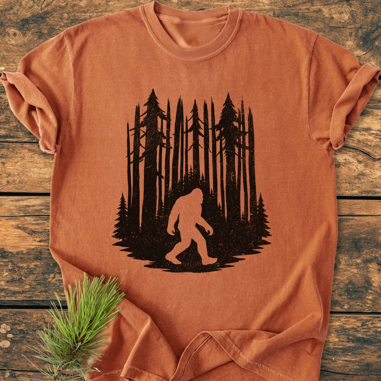 Nighttime Wanderer - Vintage Wash Tee featuring a Sasquatch silhouette in front of pine trees, displayed on a wooden surface, highlighting its relaxed fit and soft, pre-shrunk 100% ring-spun cotton fabric.