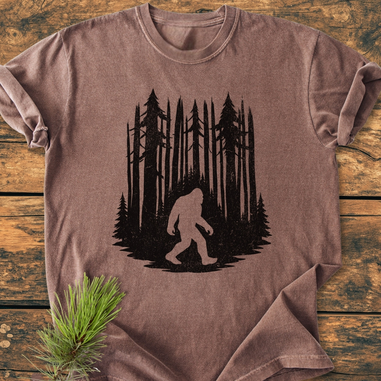 Nighttime Wanderer - Vintage Wash Tee displays a black silhouette of a pine tree forest and Bigfoot. Laid flat with a pine sprig beside it, showcasing its relaxed fit and durability.