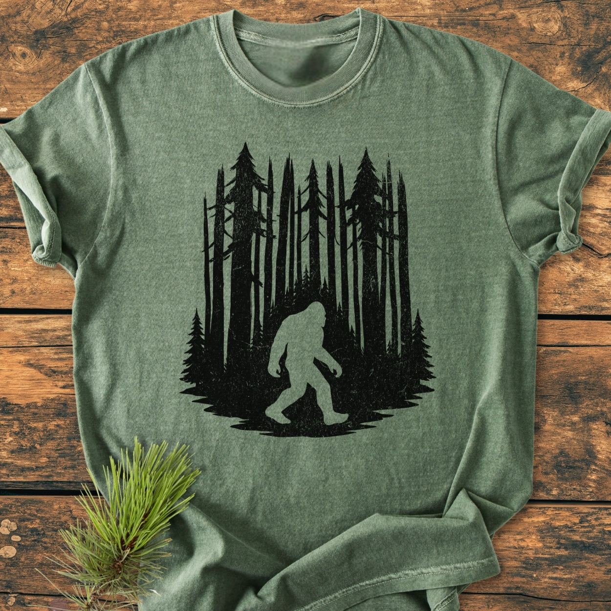 Nighttime Wanderer - Vintage Wash Tee featuring a black pine forest and Bigfoot design, laid on wood. Includes a pine sprig, highlighting its relaxed fit and durable, soft-washed fabric.