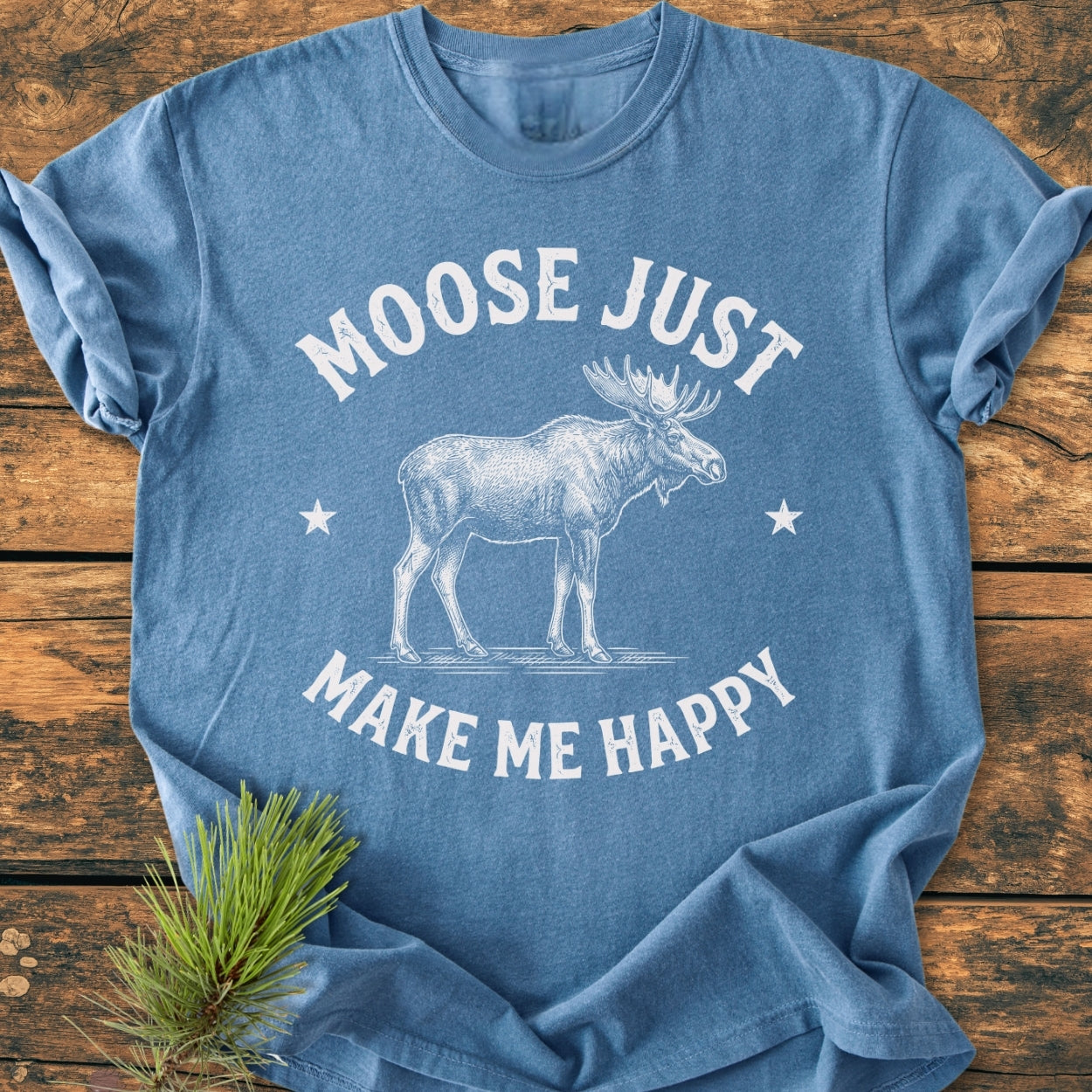 Heather-blue Moose Make Me Happy - Vintage Wash Tee with a moose sketch and stars, laid on wooden surface, featuring soft, garment-dyed fabric and relaxed fit, with pine needles beside it.
