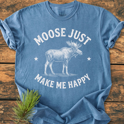 Heather-blue Moose Make Me Happy - Vintage Wash Tee with a moose sketch and stars, laid on wooden surface, featuring soft, garment-dyed fabric and relaxed fit, with pine needles beside it.