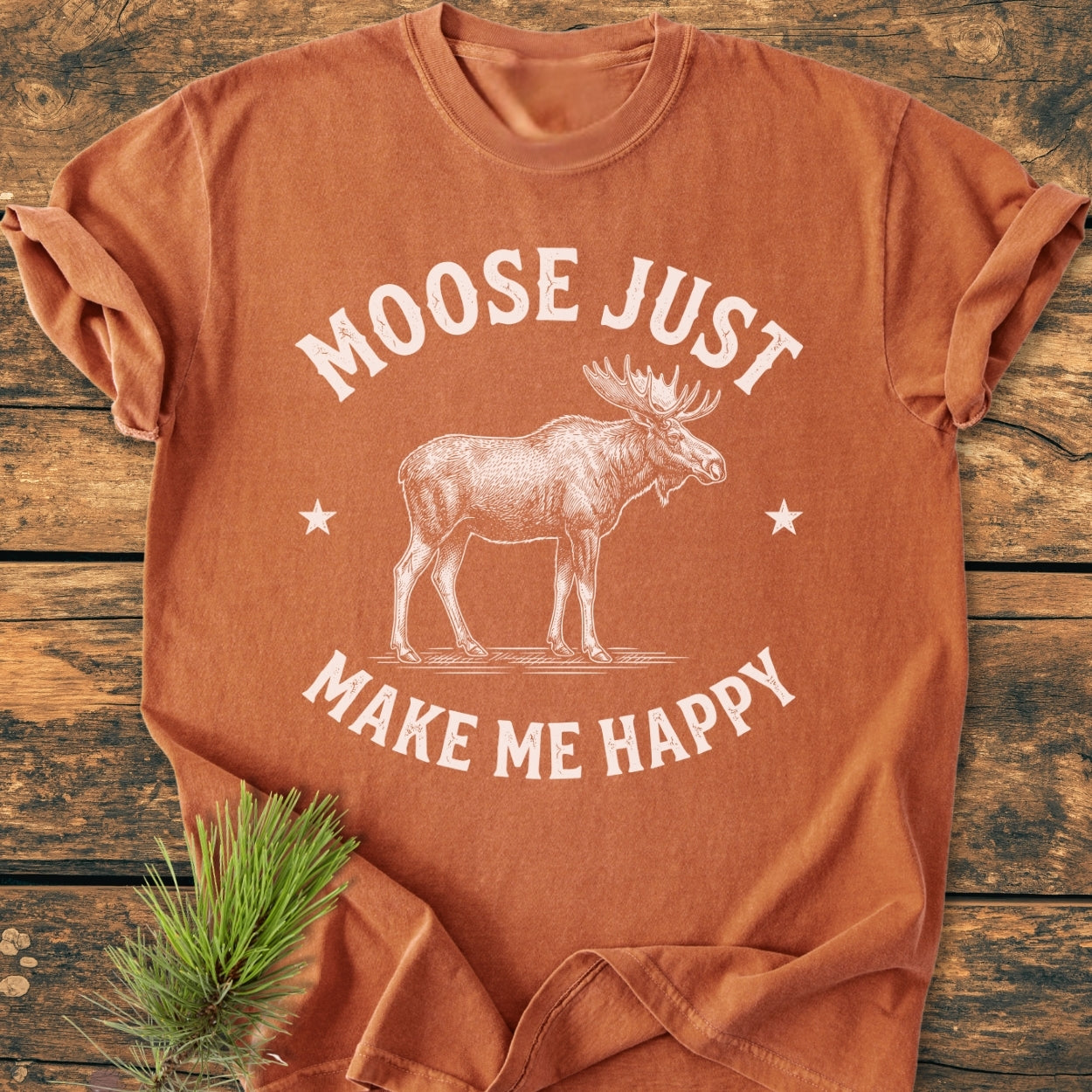 Moose Make Me Happy - Vintage Wash Tee folded on wooden surface, showcasing a moose graphic with stars. Pine sprig adds a nature touch, emphasizing the tee's rustic charm and relaxed fit.