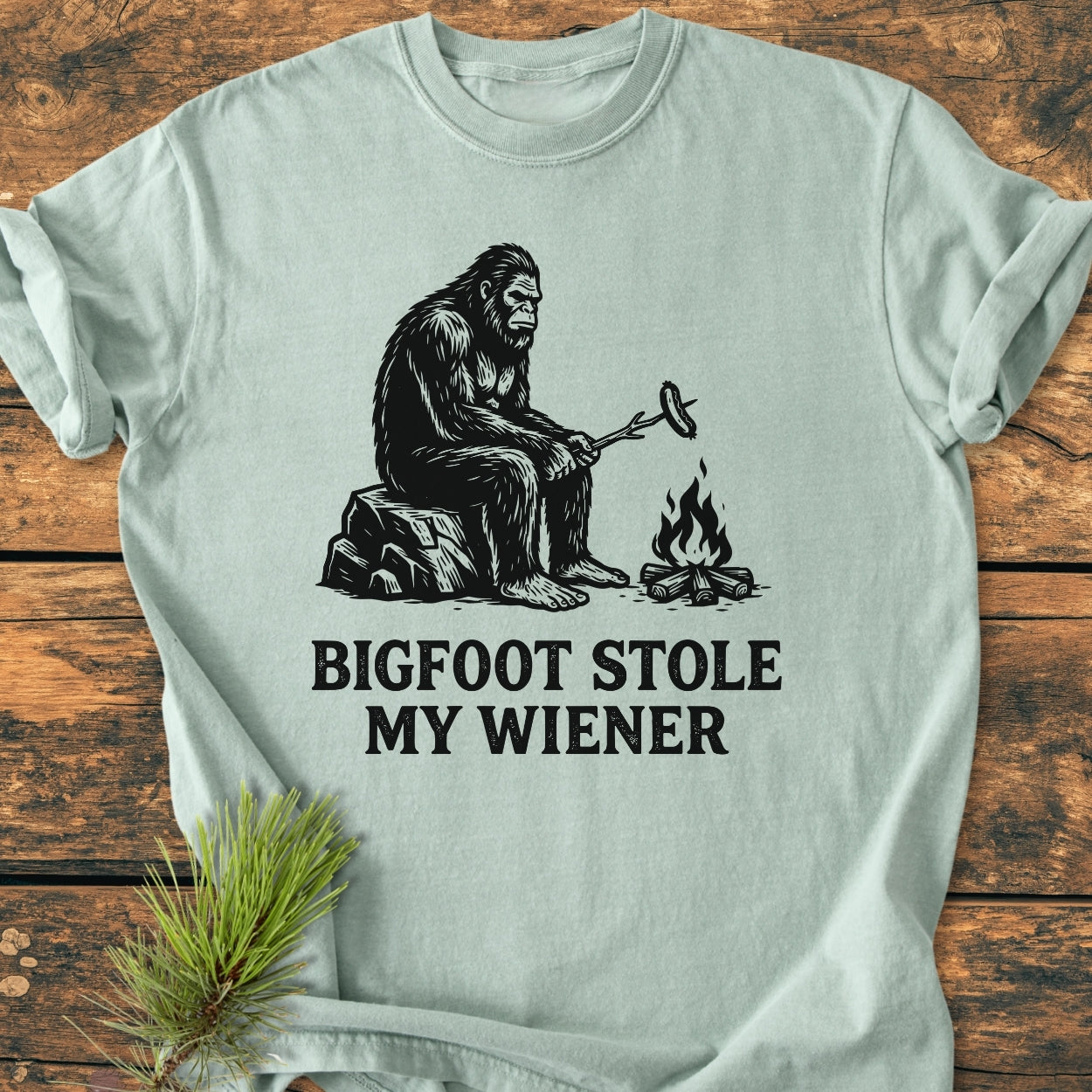 Bigfoot Stole My Wiener - Vintage Wash Tee features a cartoon Bigfoot roasting a sausage over a campfire with whimsical text below, highlighting its playful theme.