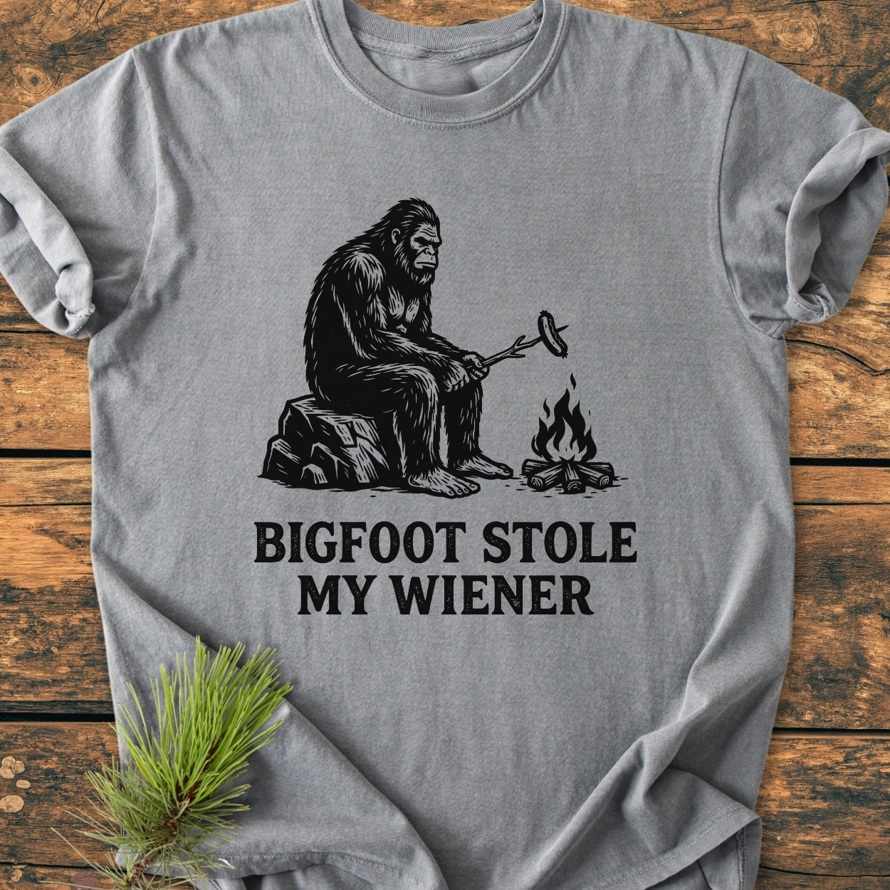 Bigfoot Stole My Wiener - Vintage Wash Tee features a black line-drawing of Bigfoot by a campfire with a hot dog, captioned humorously. The gray tee offers comfort with its relaxed fit and durable stitching.