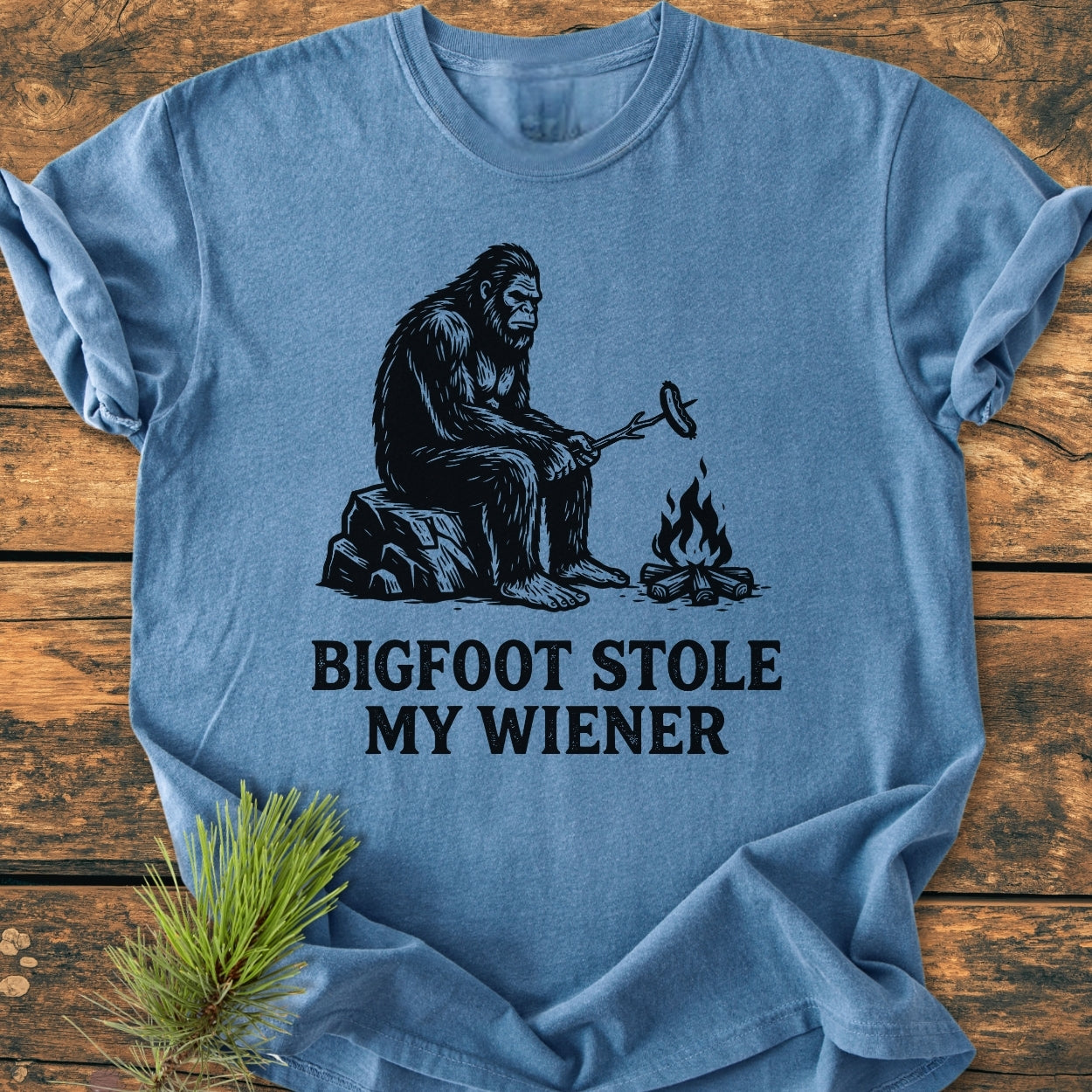 Bigfoot Stole My Wiener - Vintage Wash Tee: Light-blue shirt features cartoon Bigfoot roasting a hot dog over a campfire, captioned humorously. Laid on wood surface with decorative pine twig.