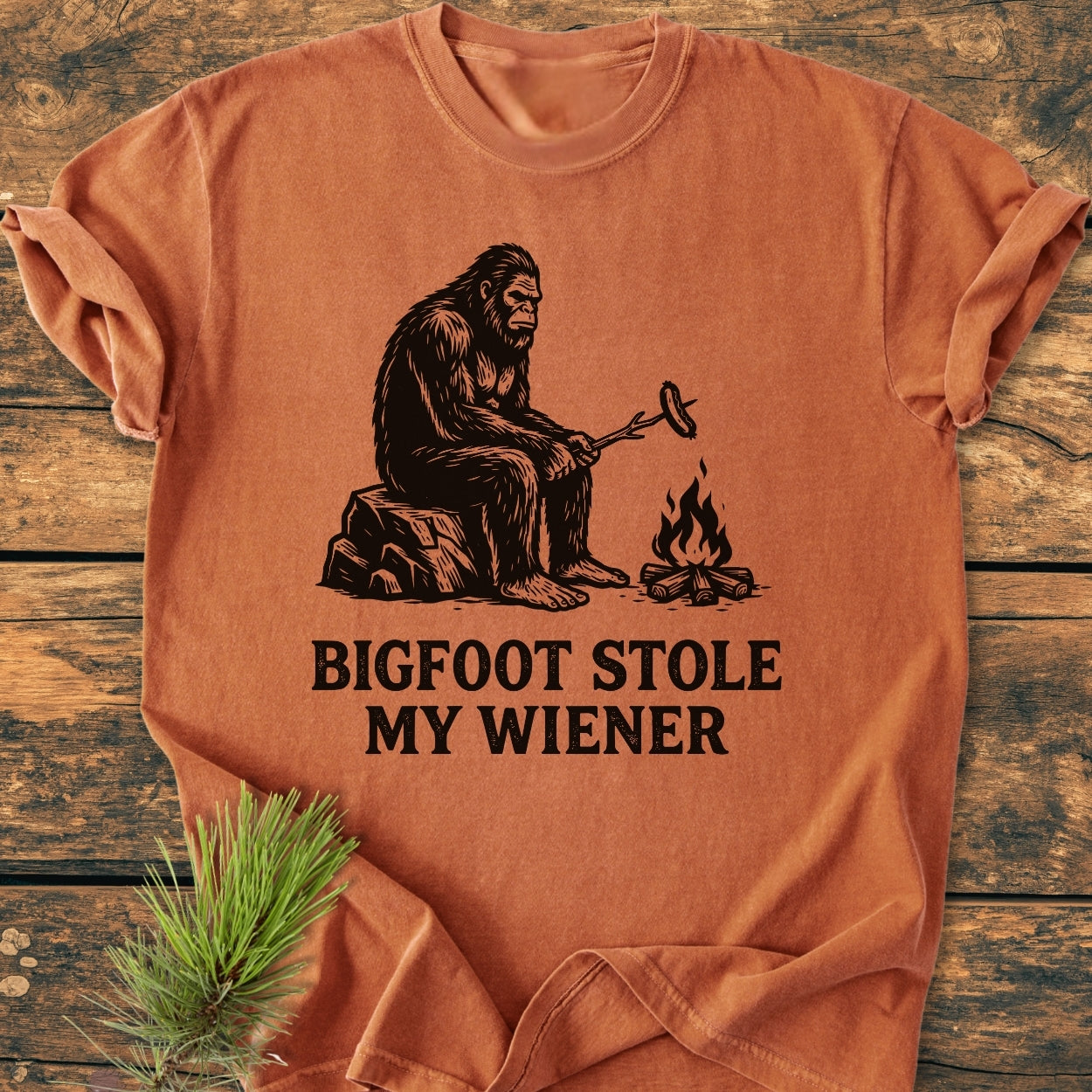 Bigfoot Stole My Wiener - Vintage Wash Tee features a black line-drawing of Bigfoot roasting a sausage over a campfire on a soft, garment-dyed T-shirt.