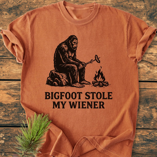 Bigfoot Stole My Wiener - Vintage Wash Tee features a black line-drawing of Bigfoot roasting a sausage over a campfire on a soft, garment-dyed T-shirt.