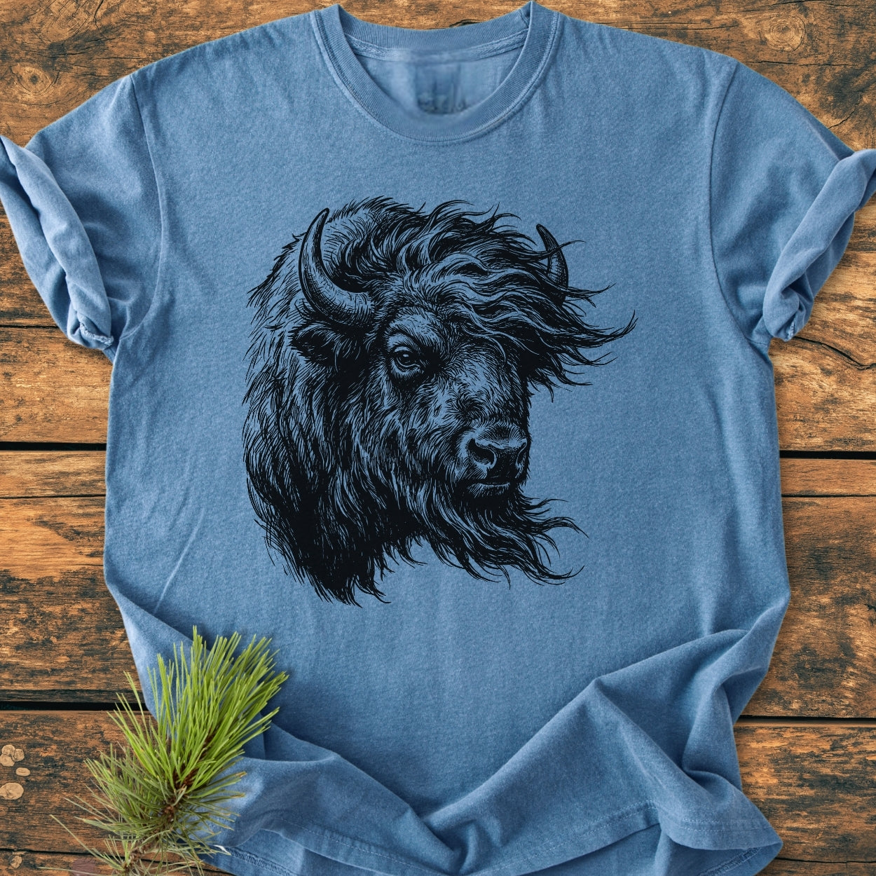 Silent Thunder - Vintage Wash Tee features a detailed black line-art bison head illustration on a light-blue cotton shirt. Laid flat on wood, it highlights comfort and durability, with pine sprig accent.