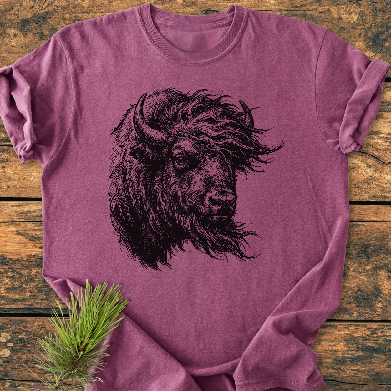Silent Thunder - Vintage Wash Tee: A heathered purple T-shirt featuring a black, hand-drawn bison head illustration, laid flat on a wooden surface with a pine sprig accentuating its outdoorsy charm.