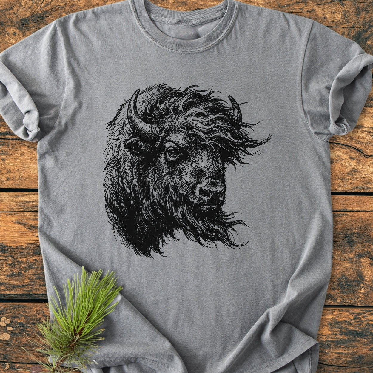 Silent Thunder - Vintage Wash Tee featuring a black bison head illustration on a heather-gray, soft-washed, short-sleeve shirt, styled with a pine sprig on a wooden surface.
