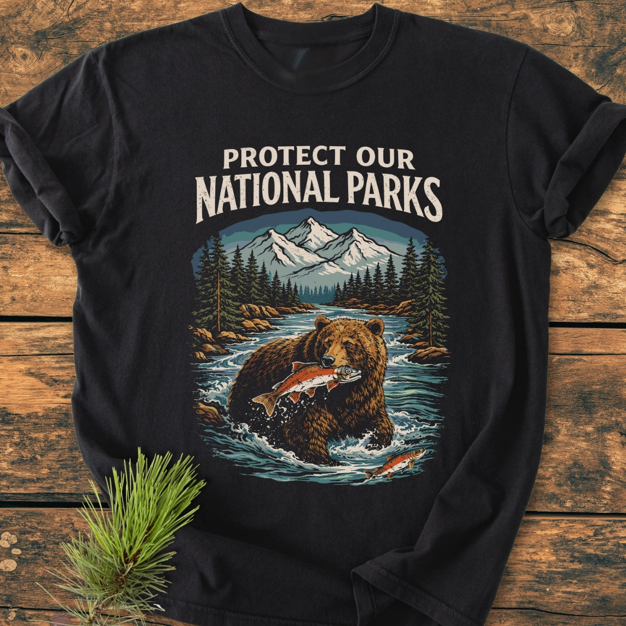 Protect Our National Parks - Vintage Wash Tee featuring a bear in a river scene with mountains, slogan above. Displayed on wood, accompanied by pine needles for a rustic touch.