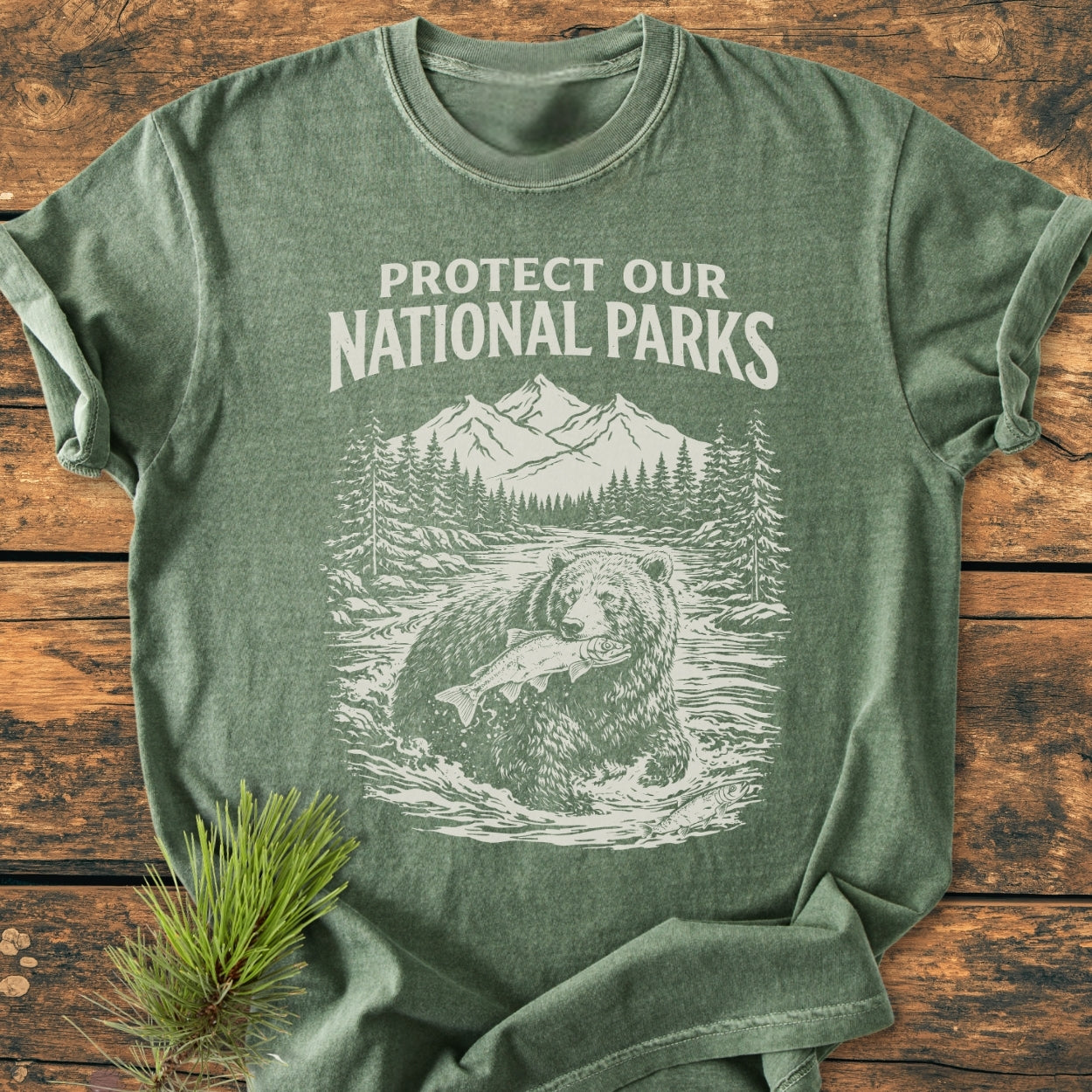 Protect Our National Parks - Vintage Wash Tee features a green crew-neck with a white slogan and outdoor scene, showcasing a bear, river, trees, and mountains, laid flat with a pine sprig.