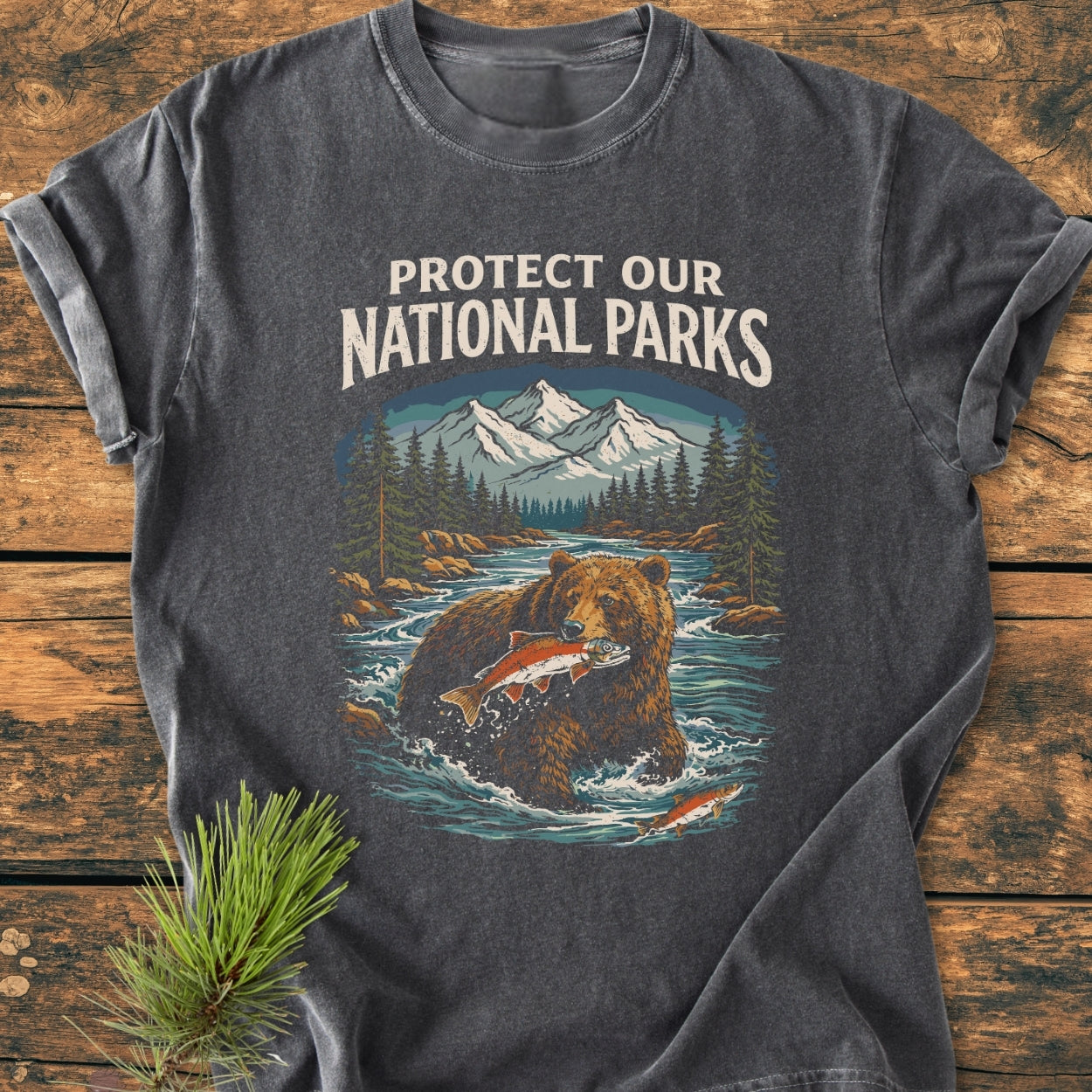 Protect Our National Parks - Vintage Wash Tee featuring a wilderness scene with mountains, evergreens, and a bear catching a fish, laid flat on wood with a pine sprig beside it.