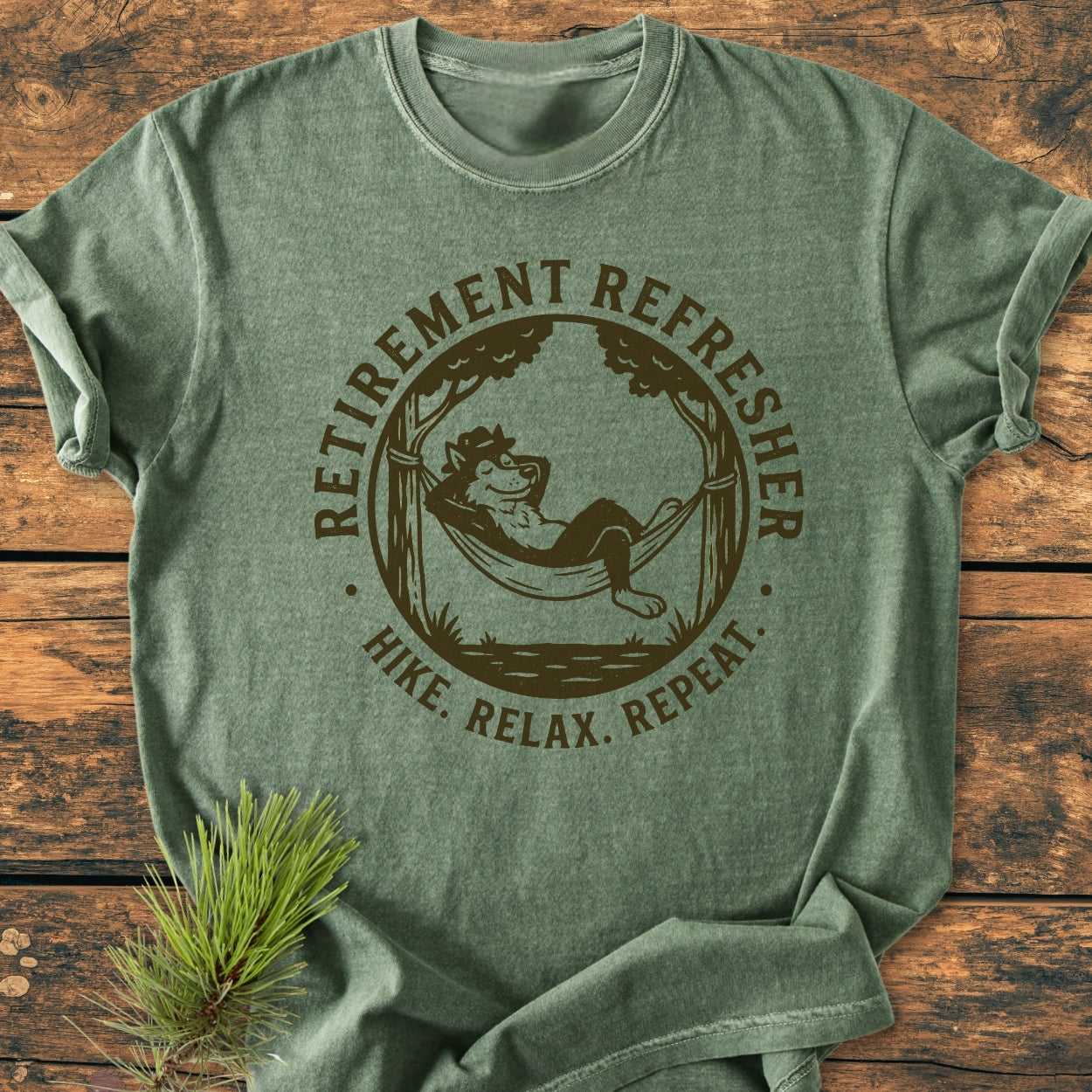 Retirement: Hike. Relax. Repeat - Vintage Wash Tee featuring a cartoon wolf lounging in a hammock, designed on a heather-green, folded, soft-washed t-shirt, displayed on a wooden surface.