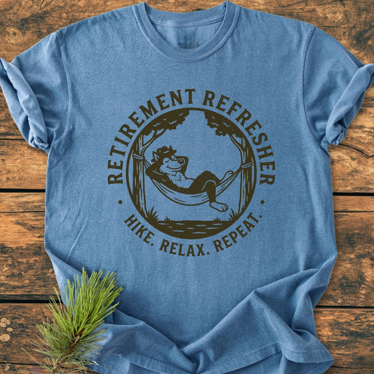 Retirement: Hike. Relax. Repeat - Vintage Wash Tee featuring a cartoonish wolf in a hammock between trees. The blue T-shirt is laid flat on wood with durable double-needle stitching and a relaxed fit.
