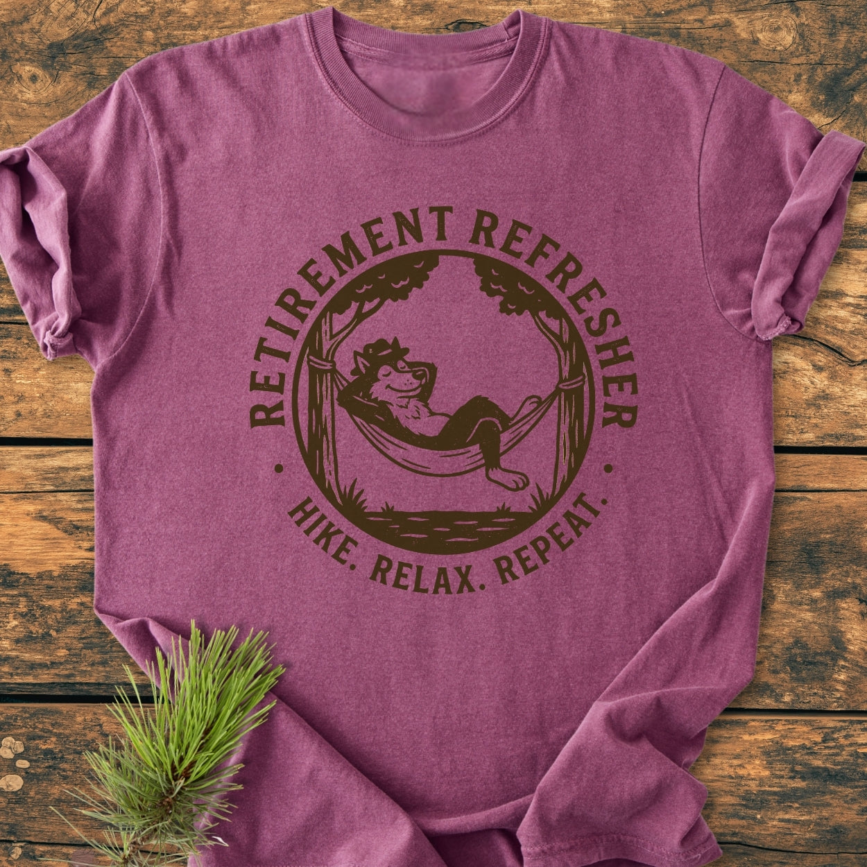 Retirement: Hike. Relax. Repeat - Vintage Wash Tee featuring a cartoon bear in a hammock with the slogan. The maroon tee lies on wood with a pine sprig for a rustic touch.