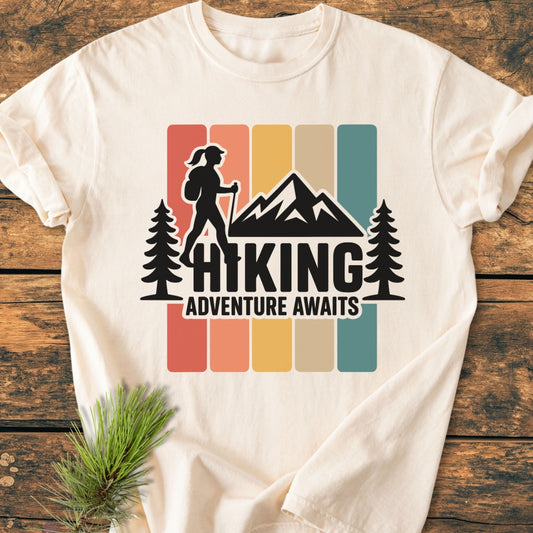 Hiking Girl - Vintage Wash Tee features a retro outdoors graphic with a hiker silhouette and slogan, laid on a wooden surface with a pine sprig beside it, highlighting its adventure theme.